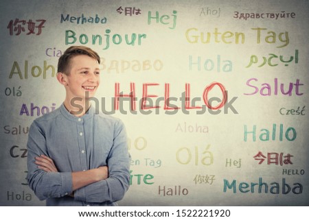 Positive boy adolescent keeps arms crossed looking aside tooth smile over grey wall background written with the word hello in different languages and colors. Learning opportunity for young students.