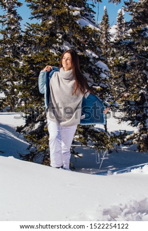 Portrait of a young beautiful brunette woman with blue eyes and freckles on face in winter snowy mountain landscape. Beautiful girl in the winter outdoors dreesed in sweater and blue jacket.