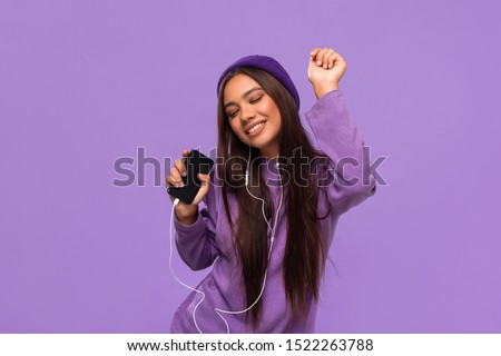 Pretty african-american girl in a hat and sweater listening to music on a mobile phone with wired headphones and dancing isolated over purple background. Enjoying life.