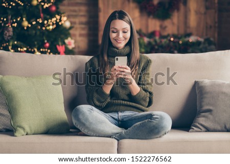 Full body photo of positive festive girl sit on divan crossing her legs using cellphone communicating enjoy christmas celebration in house full of newyear decoration lights indoors