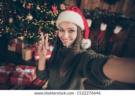 Close up photo of positive newyear blogger brunette hair girl in knitted pullover take selfie make v-signs on christmas evergreen tree with many gift boxes wear santa claus cap in house indoors