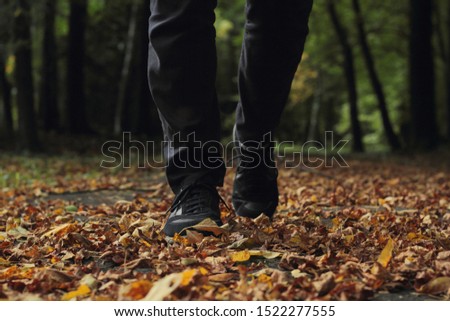 Closeup of man's legs walking in dry leaves on a pavement with trees in the background.