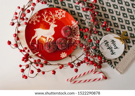Christmas and new year flat lay. Red plate with a picture of a deer, red apples, candy cane, Christmas tree toy and branches with red berries on a white background. The view from the top