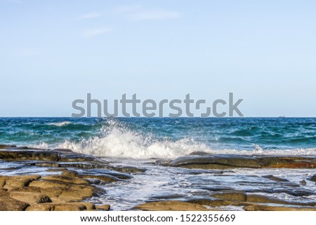 Waves crashing against flat lava rocks jutting into the ocean with freight ships dotting the horizon