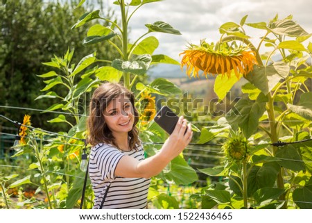 A pretty young millennial brunette woman taking a selfies beside a sunflower field.