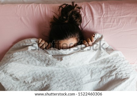 Playful young woman hiding face under blanket while lying in cozy bed, pretty curious girl feeling shy peeking from duvet, covering with white sheet, head shot close up. Top view Royalty-Free Stock Photo #1522460306