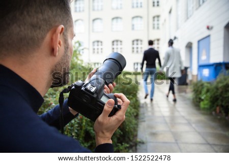 Young Man Paparazzi Photographer Capturing A Photo Suspiciously Of Couple Walking Together Using A Camera Royalty-Free Stock Photo #1522522478