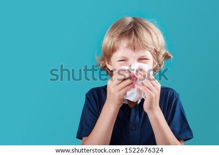 Little school boy with blonde hair sneezing to handkerchief and having runny nose. Flue, cold, allergy and disease concept Royalty-Free Stock Photo #1522676324