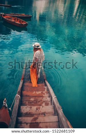 Girl in a straw hat on the background of the turquoise lake with wooden boats in mountains. Safari style blonde woman back view. Dolomites Alps, lago di Braies, Italy