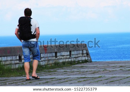 A man with a black backpack on the roof of an old building on a background of the sea. The view from the back.