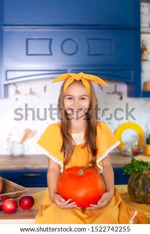 Little girl holds big pumpkin in kitchen at home. Harvesting. Healthy nutrition, vegetarianism, vitamins, vegetables. Cute child getting ready for Halloween and having fun with pumpkins in the kitchen