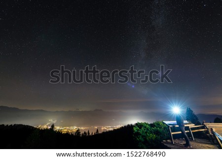 Man with luminous headlamp looks on the starry sky over the nocturnally lit Rottach-Egern at the Lake Tegernsee, Bavaria, Germany