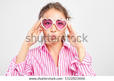 Cool Asian girl in heart-shaped glasses fools around and poses on a white background, pulls her lips into a straw, pretends to kiss