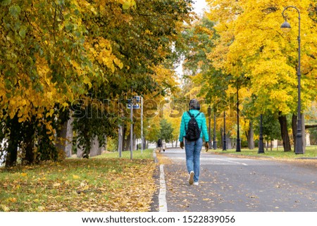 A picture of a lonely girl in a short warm jacket, blue jeans and a backpack. Walk along an asphalt road covered with fallen yellow leaves in an autumn park among the trees. back view