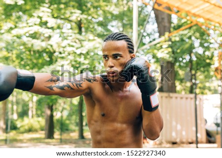 Sweated bare-chested sportsman with boxing gloves exercising outdoors