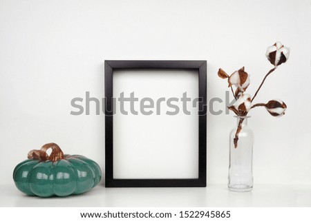 Mock up black frame with pumpkin decor and cotton flowers on a shelf or desk. Autumn concept. Portrait frame against a white wall.