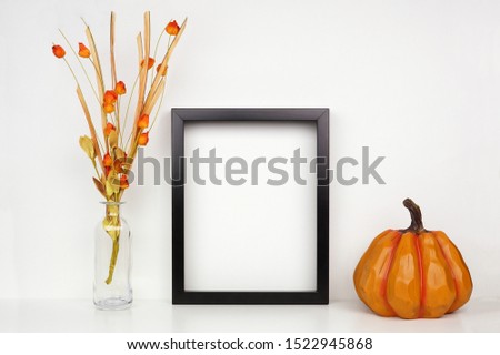 Mock up black frame with fall branches and pumpkin decor on a shelf or desk. Autumn concept. Portrait frame against a white wall.