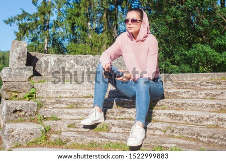 A young woman in a pink hoodie and sunglasses sitting on a stone bench and operated by remote control drone flight in nature. Full-length photo. Blurred background. Drone in the foreground.