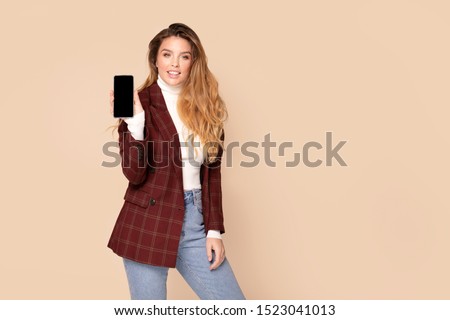 Smiling young woman with freckles and long wavy hair holding mobile phone with blank empty screen isolated on studio background. People lifestyle concept. Autumn, winter style.