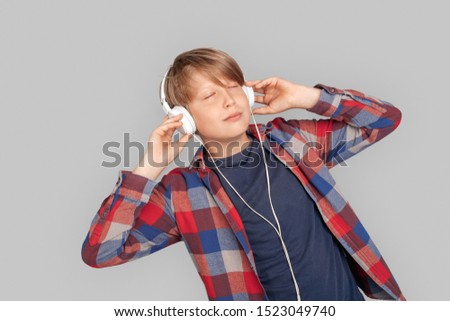 Young boy wearing headphones standing isolated on grey background listening to music closed eyes concentrated