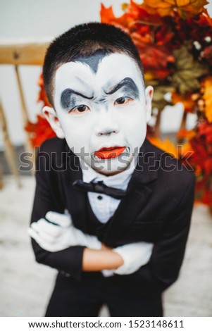 Asian boy with dracula face painting and full costume. Outdoor photo shoot. Vintage furniture and decor. Fall leaves wreath. Stylish young kid ready for trick or treat. Classy suit for halloween.