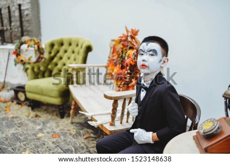 Asian boy with dracula face painting and full costume. Outdoor photo shoot. Vintage furniture and decor. Fall leaves wreath. Stylish young kid ready for trick or treat. Classy suit for halloween.