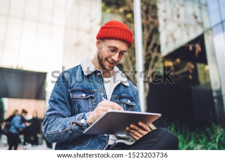 From below of hipster man using modern tablet with stylus while creating new project and sitting on street near glass skyscraper