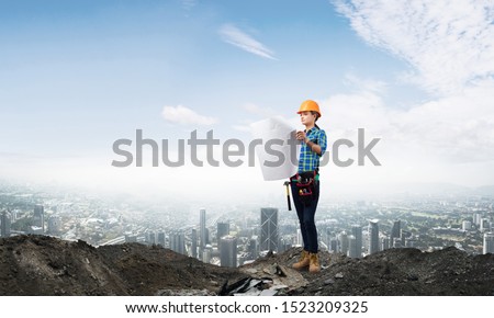Female engineer in hardhat standing with technical blueprint on construction site. Young woman architect in workwear inspecting construction on background of cityscape. Building project management.