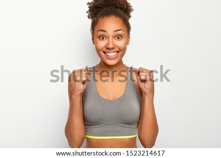 Photo of mixed race athlete female clenches fists with joy, celebrates victory of marathon, smiles broadly, dressed in grey top, smiles broadly, enjoys triumph, poses indoor against white wall