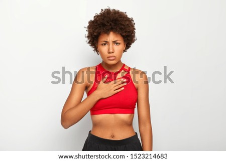 Displeased dark skinned female feels acute pain in chest, breathes hardly, wears sport clothes, looks upset, has Afro hairstyle, isolated over white background. People, negative feelings and sickness