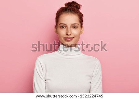 Isolated shot of pleasant looking young woman wears white turtleneck, has hair bun, looks with calm facial expressions, poses against pink background. People, natural beauty and youth concept