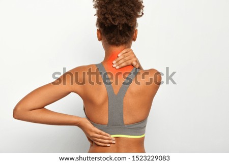 Faceless woman with dark skin suffers from nape pain, holds hand on neck with red spot, has problems with health, spine disease, wears sport bra, isolated over white background. Pain syndroms
