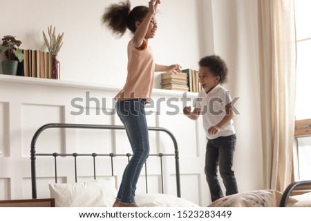 Overjoyed african american happy girl and preschooler boy jumping, having fun, playing on bed in bedroom. Cute smiling children brother and sister enjoy leisure weekend morning activities.