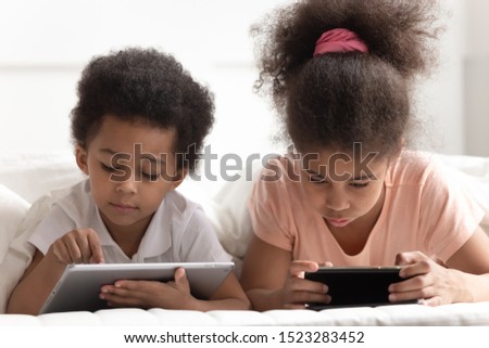 Concentrated cute little boy and girl laying on bed, using gadgets, reading, studying, playing games, watching cartoons or movies, scrolling social networks. Tech addicted children using devices.