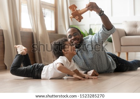 Happy smiling loving black single father in eyeglasses holding wooden plane in air, playing with little mixed race cute curly funny child son, lying on heated floor, spending weekend leisure time. Royalty-Free Stock Photo #1523285270