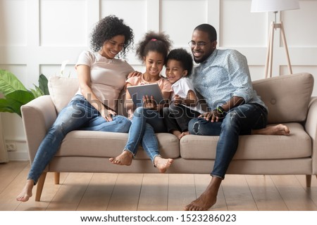 Happy parents with smiling kids sitting on sofa, using tablet looking at screen, watching cartoons online, funny videos together, little girl showing family photos on gadget, making order, ecommerce