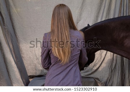 picture long-haired girl standing with her back in a jacket next to his horse gray background
