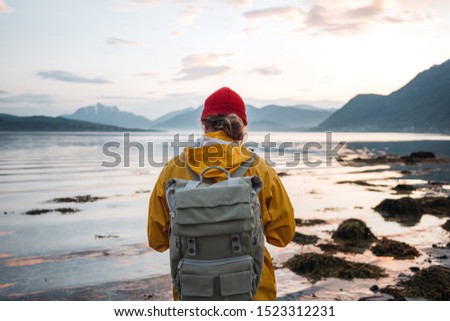 Back view of male tourist with rucksack standing on coast in front of great mountain massif while journey.  Man traveler wearing yellow jacket with backpack explore scandinavia nature. Wanderlust