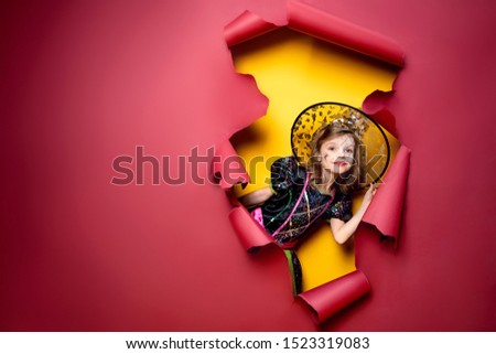 Happy Halloween. Laughing funny child girl in a witch costume of halloween looking, smiling and scares through a hole of red, yellow paper background. Copyspace