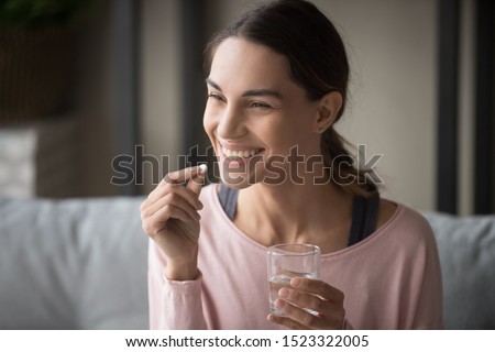 Smiling healthy young woman holding pill glass of water sit on sofa at home, positive lady take daily medicine antioxidant diet vitamin supplements for beauty skin hair health care medicament concept Royalty-Free Stock Photo #1523322005