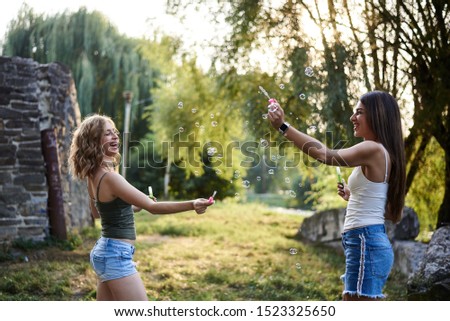 Two sisters, blowing soap bubbles at each other in park, laughing, smiling, Young pretty girls, wearing jeans shorts, green beige tops, having fun outside. Three-quarter portrait of two girlfriends.