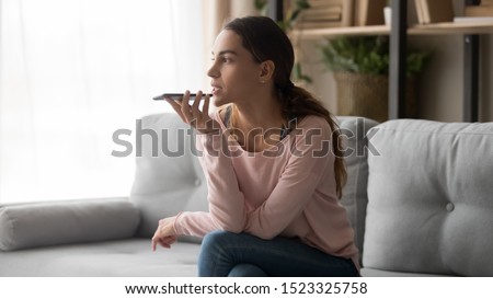 Serious young woman holding phone using mobile voice recognition or digital assistance app speak activate virtual assistant, record message or talk on speakerphone on smartphone sit on sofa at home 