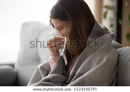 Ill upset young woman sitting on sofa covered with blanket freezing blowing running nose got fever caught cold sneezing in tissue, sick girl having influenza symptoms coughing at home, flu concept Royalty-Free Stock Photo #1523330795