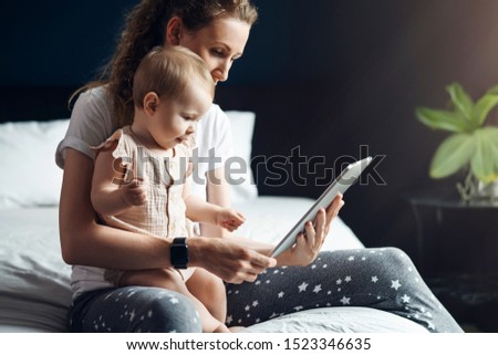 Nine-month-old baby girl with mom are sitting on the bed and looking at the screen of a digital tablet. Child development using modern technology. The child is talking on a video call. Online learning