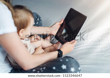 Back view.Close-up of tablet computer in hands of mother sitting with baby. Toddler girl sitting with mom on her lap, looking at screen of smartphone, watching cartoons, talking on video call.