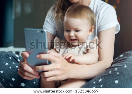 Nine-month-old baby girl sits with her mother on bed and looks on smartphone cartoons, plays game. Child is talking to her grandmother via video link. Mom shows her daughter pictures on phone screen.