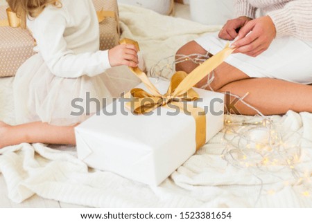 Mom and little daughter are holding Christmas and New Year presents while sitting on the floor against the background of white Christmas gifts with gold ribbons and bows.