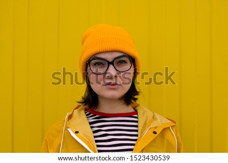 candid portrait of caucasian woman wearing yellow hat,glasses and raincoat standing against yellow wall and looking at the camera 