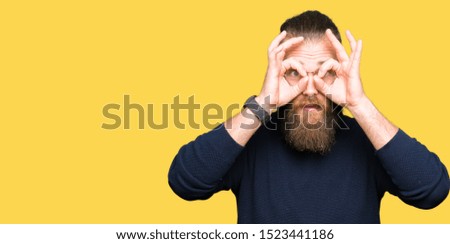 Young blond man wearing glasses and turtleneck sweater doing ok gesture like binoculars sticking tongue out, eyes looking through fingers. Crazy expression.