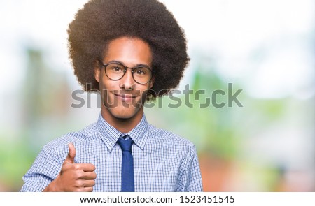 Young african american business man with afro hair wearing glasses doing happy thumbs up gesture with hand. Approving expression looking at the camera showing success.
